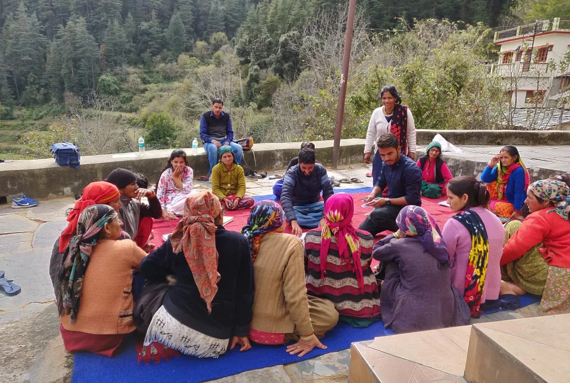 Village women and men sit together on a blue mat for a meeting