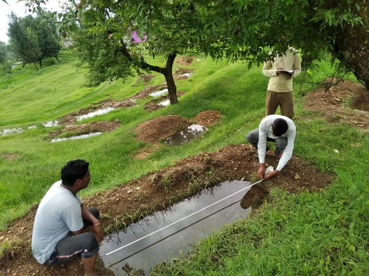 Men measure a water-filled trench on a green hillside